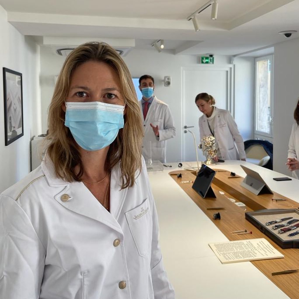 Woman in Jaeger-LeCoultre lab coat and mask, with watchmakers and tools at a table.