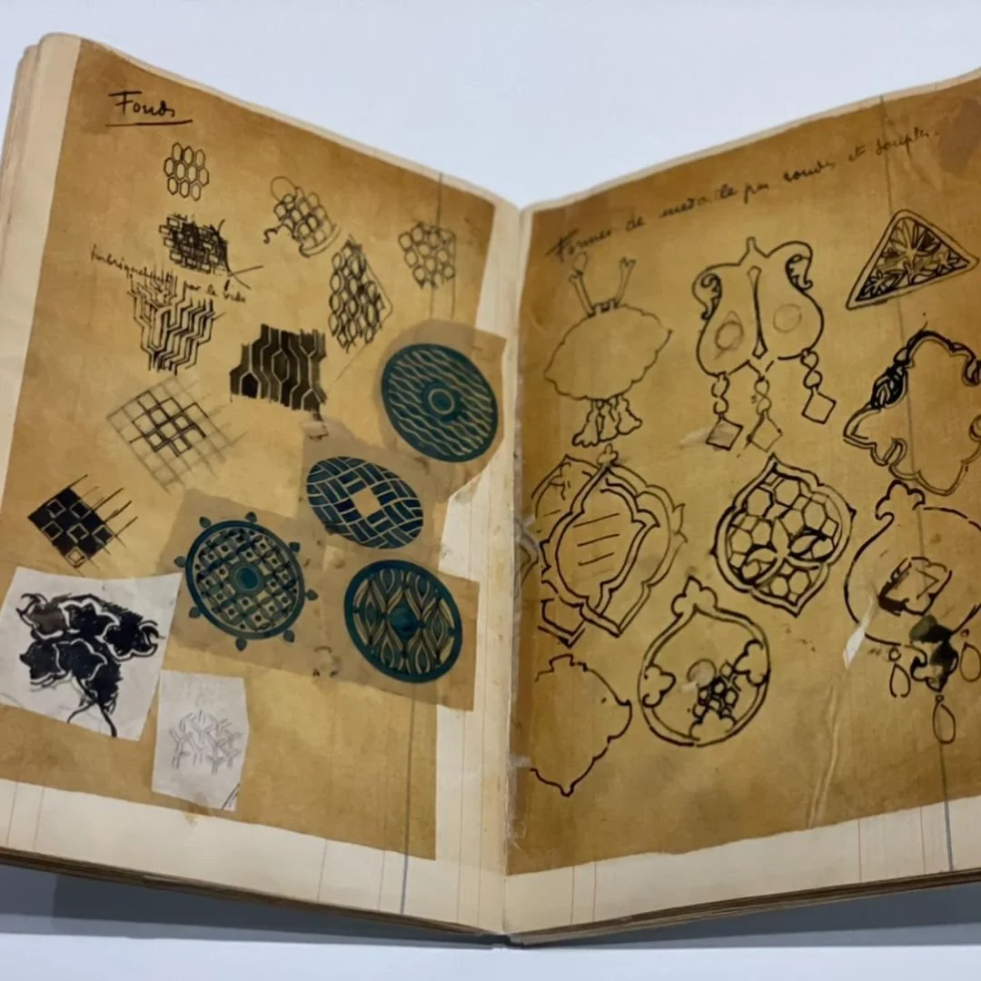 Hand-drawn jewellery designs and geometric patterns fill an open, aged sketchbook.
