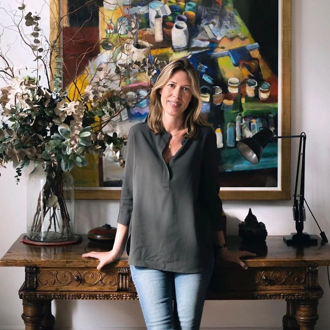 Francesca Cartier Brickell smiling, wearing a gold cross necklace, standing by a carved wooden table.