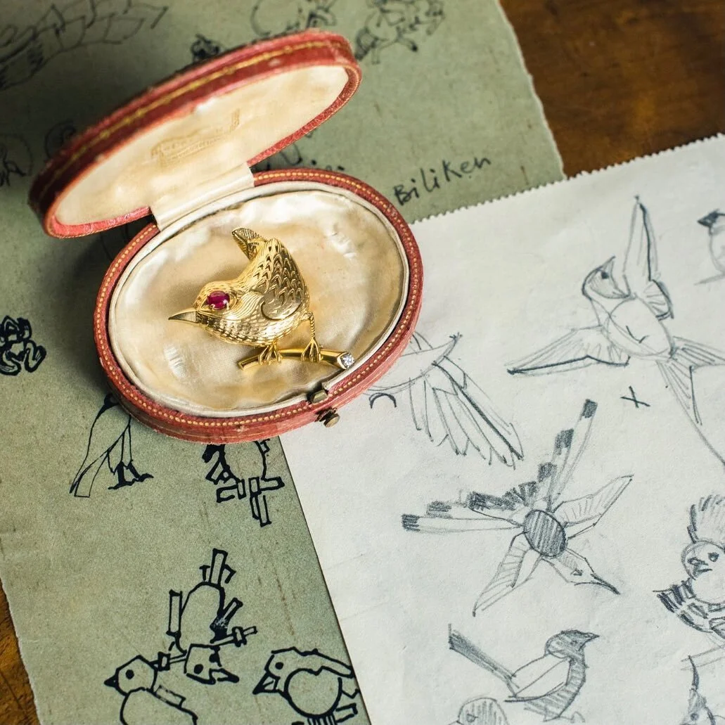 Gold bird brooch with ruby eye and diamond, in red box, surrounded by bird sketches.