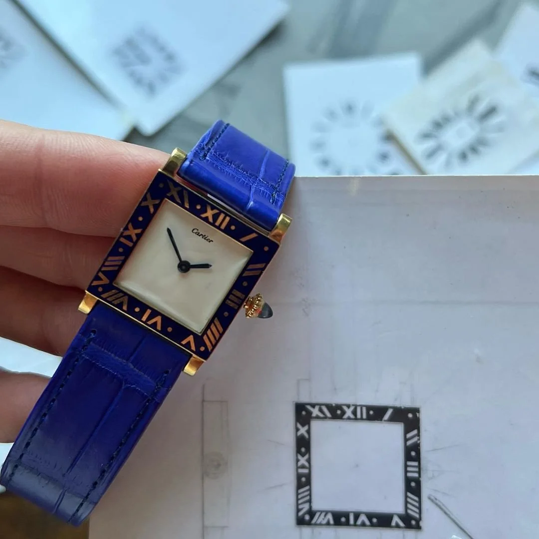 Vintage gold Cartier Tank watch with blue enamel bezel, cream dial, and blue alligator strap.