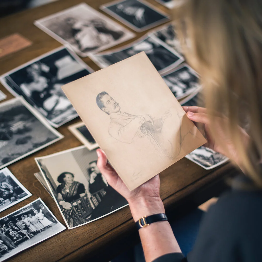 Woman's hands, wearing a Cartier watch, hold a drawing of a man, surrounded by old photos.