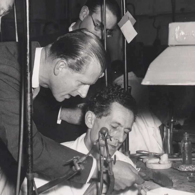 A man in a suit leans over to watch a jeweller at his workshop bench.