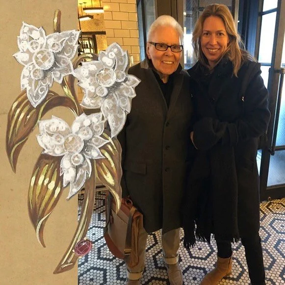 Two women smile beside a large illustration of a diamond floral brooch with gold leaves.