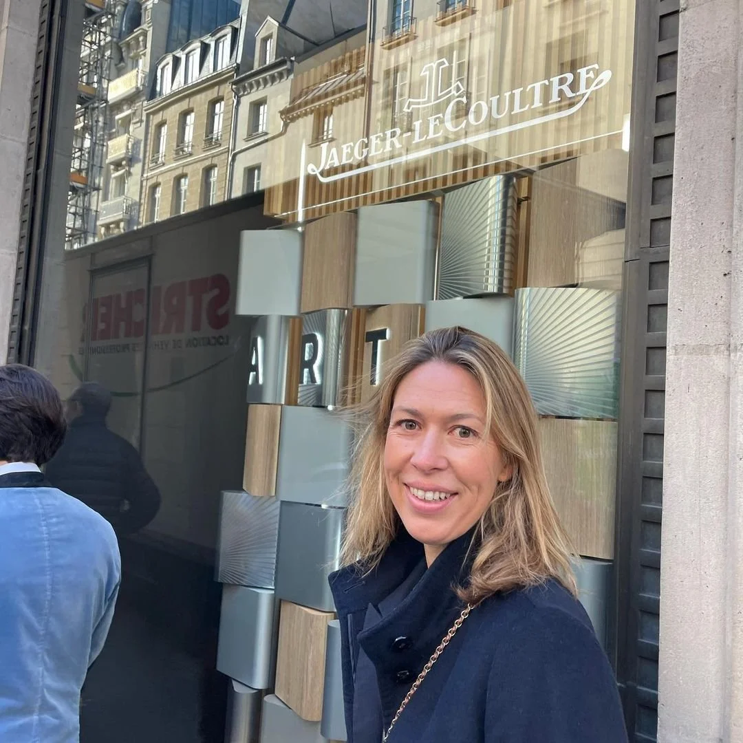 Francesca Cartier Brickell smiles before a Jaeger-LeCoultre store window reflecting buildings.
