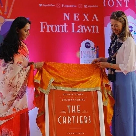 Two women unveil a large orange book titled 'THE CARTIERS' at a literature festival.