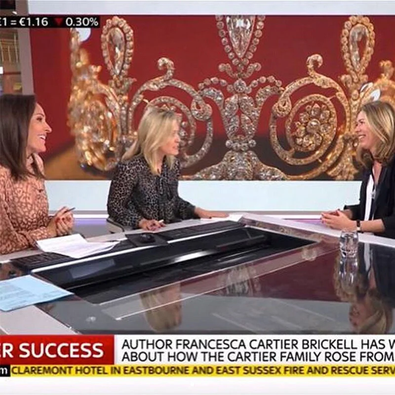 Three women conversing at a desk, with ornate diamond and gold jewellery on a red screen.